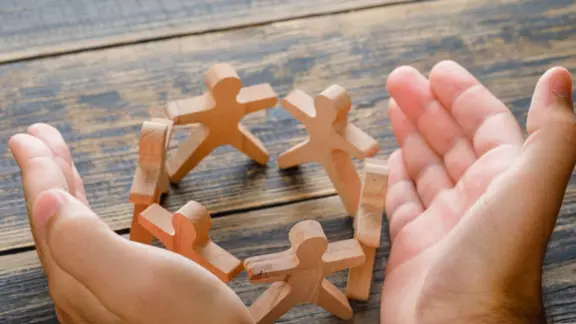 houten mannetjes staan in een kring en handen raken elkaar. 2 volwassen handen omkaderen het houten werk