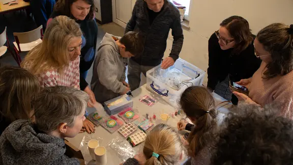 Een tiental deelnemers staat rond een tafel te kijken naar hoe je een epoxy sleutelhanger maakt