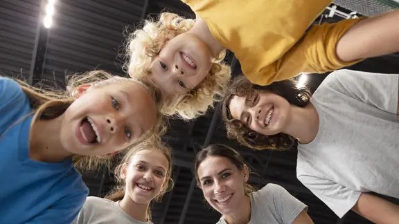 5 jongeren staan in een kring met hun hoofden bij elkaar en kijken naar beneden, naar de camera