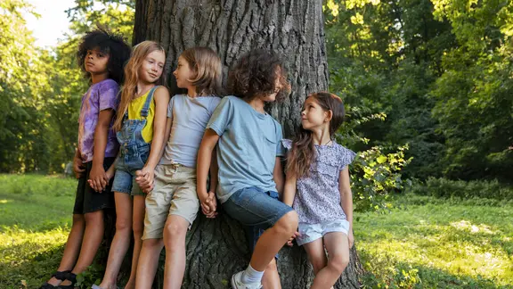 5 kinderen staan met hun rug tegen een dikke boomstronk